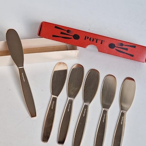 May include: Set of six gold-toned butter spreaders with a matching spreader and a red box. The box has the word "POTT" printed on it, along with utensil illustrations. The spreaders are arranged on a white surface.