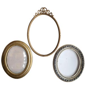 Op de afbeelding: Drie antieke fotolijsten in goud- en zilverkleuren. De lijsten zijn ovaal en rond, met sierlijke details. Eén lijst heeft een decoratieve strik aan de bovenkant. De lijsten zijn leeg, klaar voor foto's of kunstwerken.