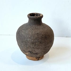 Peut inclure: Un petit vase rond en céramique brun foncé avec un col étroit et un bord légèrement évasé. Le vase a une surface rugueuse et texturée et repose sur une petite base circulaire. La forme générale rappelle un ancien récipient.