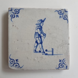 Op de afbeelding: Vierkante, gebroken witte tegel met een blauw-witte illustratie van een persoon die een spel speelt met een stok en een bal. De tegel heeft blauwe decoratieve hoekontwerpen. De persoon draagt een hoed en een korte broek. De tegel heeft scheuren.