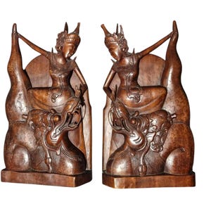 Op de afbeelding: Twee houten sculpturen van zittende figuren met sierlijke hoofdtooien en vloeiende kleding. Elke figuur zit bovenop een gedetailleerde draak, met uitgestrekte armen. De sculpturen hebben een warme bruine kleur en lijken uit één stuk hout gesneden.