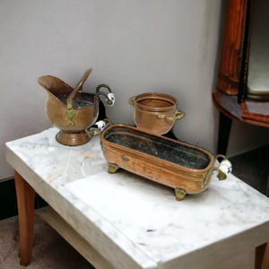 Op de afbeelding: Een assortiment antieke koperen containers, waaronder een kleine pot, een kan en een rechthoekige plantenbak. De koperen items staan op een tafel met een marmeren blad en houten poten. De plantenbak heeft decoratieve handvatten.