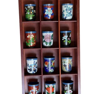 Pode incluir: Uma prateleira de exposição retangular de madeira com doze compartimentos, cada um contendo um copo cilíndrico decorado com padrões florais cloisonné coloridos. Os copos apresentam vários desenhos, incluindo crisântemos, peónias e flores de ameixeira.
