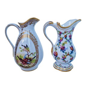 Puede incluir: Dos jarras antiguas de porcelana. Una presenta una escena de pájaros dentro de un óvalo con ribete dorado, la otra está adornada con un estampado floral. Ambas tienen detalles dorados en las asas y los bordes. Las jarras son blancas con acabado brillante.