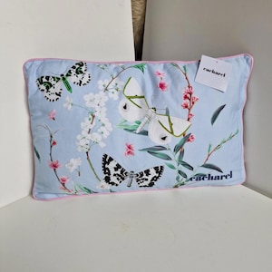 Op de afbeelding: Een rechthoekig lichtblauw kussen met een roze rand, met een bloemen- en vlinderontwerp. Het kussen heeft een label met de tekst "caharel". De vlinders zijn wit, zwart en groen, met roze en witte bloemaccenten.
