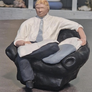 Op de afbeelding: Een beeldje van een persoon die in een zwarte stoel zit. De figuur draagt een wit overhemd, een lichtblauwe stropdas en een donkerblauwe broek. De figuur heeft blond haar en houdt een wit kussen en een lichtblauw kussen vast.