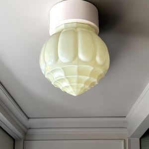Op de afbeelding: Een plafondlamp met een lichtgele, gegoten glazen kap met een getextureerd, gesegmenteerd patroon. De kap is bevestigd aan een eenvoudige, effen witte ronde basis die vlak tegen het platte plafondoppervlak aanligt.