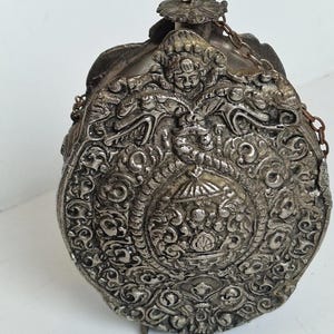 May include: An antique silver-coloured flask with intricate relief carvings. The flask has a central medallion with floral and geometric patterns, surrounded by figures and scrollwork. A small chain is attached to the top.