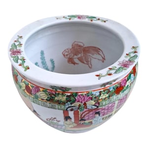 Puede incluir: Una pecera de cerámica blanca con un diseño floral y paisajístico detallado. El cuenco presenta un pez dorado pintado y adornos decorativos con flores rosas y hojas verdes. El exterior muestra una escena colorida con figuras y patrones intrincados.