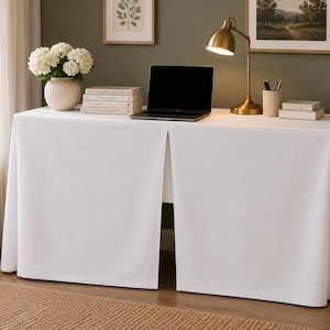 May include: A rectangular table covered with a floor-length white fabric tablecloth featuring a central inverted pleat. The fabric drapes over the sides and front, creating a clean, structured appearance that conceals the table legs and frame.
