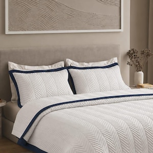 May include: A white bedspread and two matching pillow shams featuring a textured herringbone pattern and a solid navy blue border. The bedding set is displayed on a bed with a neutral-coloured headboard, providing a clean and structured appearance.
