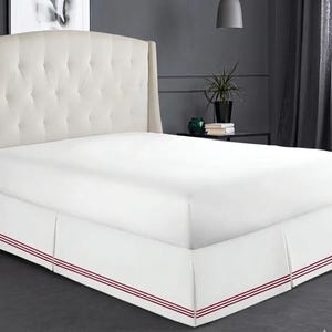 May include: A white bed with a tufted headboard and a white bed skirt. The bed skirt has three thin red stripes near the bottom. The bed is in a room with a dark gray wall and a dark gray floor.