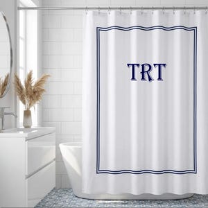 以下が含まれることがあります： ネイビーブルーの縁取りとネイビーブルーの文字「TRT」が入った白いシャワーカーテン。白い洗面化粧台、鏡、バスタブのあるバスルームにカーテンが吊るされています。