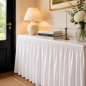 May include: A white console table draped in a floor-length white fabric skirt with gathered pleats. The table surface holds a cream ceramic lamp with a pleated shade, a stack of three books titled Live Beautifully, This Is Home, and The Kinfolk Home.