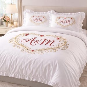 May include: White bedding set with a large embroidered heart design in gold and red, featuring the letters "A&M". Two matching pillowcases with ruffled edges and similar heart embroidery. The bed is in a bedroom setting.