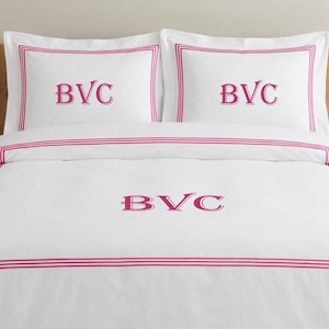 May include: White bedding set featuring a pink border and embroidered initials "BVC". The set includes two pillowcases and a duvet cover. The duvet cover and pillowcases have a simple, modern design.