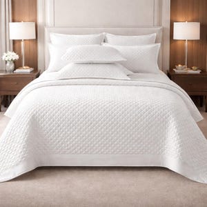 Puede incluir: Una cama blanca con un edredón acolchado y almohadas a juego. La cama tiene un cabecero blanco, dos mesitas de noche con lámparas y una alfombra de color neutro.