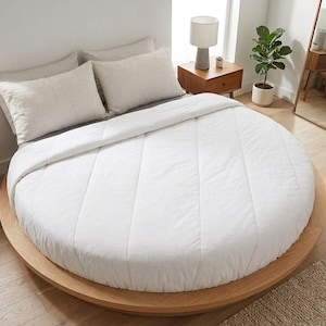 May include: A round bed with a light wood frame and a white comforter. Two beige pillows rest against the headboard. A small side table with a lamp and a potted plant are visible in the background. The bed is the focal point of the room.