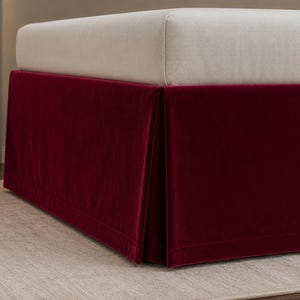 May include: A deep burgundy velvet bed skirt featuring a tailored design with clean lines and a smooth, matte finish. The fabric drapes from the base of a light-colored mattress, creating a structured look that covers the lower frame of the bed.