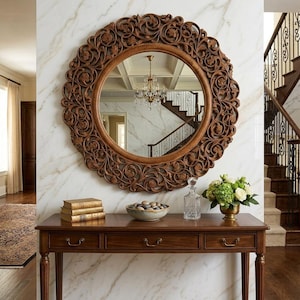 May include: A round wall mirror with a dark brown wooden frame featuring intricate carved floral and scrollwork patterns. The mirror is mounted on a white marble wall above a rectangular wooden console table with three drawers and brass pulls.