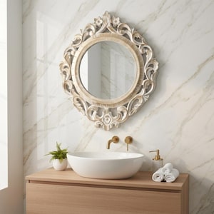 May include: A round wall mirror with an ornate, carved light-wood frame featuring intricate scrollwork and floral details. The mirror is mounted above a white oval vessel sink, a gold faucet, a small potted green plant, and white towels.