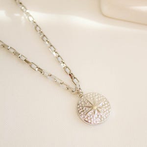 Puede incluir: Un collar plateado con un colgante circular. El colgante presenta un diseño de estrella y una superficie texturizada. La cadena está hecha de eslabones rectangulares. El collar se muestra sobre un fondo blanco.