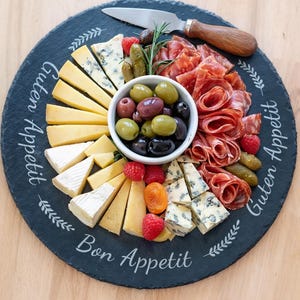 Könnte beinhalten: Ein runder schwarzer Servierteller aus Schiefer mit einer Auswahl an Käse, Oliven, Salami und frischen Beeren. Ein kleines Messer liegt auf dem Teller. Die Worte "Guten Appetit" und "Bon Appetit" sind auf dem Teller geschrieben.