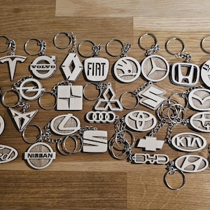 May include: A collection of wooden keychains featuring various car brand logos, including Ford, Tesla, and Jeep. Each keychain has a silver ring. The keychains are arranged on a wooden surface.
