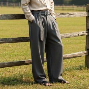 Puede incluir: Un hombre viste una camisa beige abotonada y pantalones grises de pernera ancha con cinturón a juego. El atuendo es informal y elegante. El fondo muestra una valla de madera y un campo de hierba.