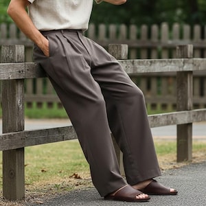 May include: A light beige short-sleeved shirt with a mandarin collar and dark brown trousers. The trousers have a pleated front and a wide leg. The person is wearing brown leather sandals. The outfit is casual and relaxed.