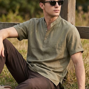 May include: An olive green short-sleeved shirt with a mandarin collar and a partial button-down placket, paired with dark brown trousers and black sunglasses. The shirt features a textured fabric and cuffed sleeves, worn in a casual setting.