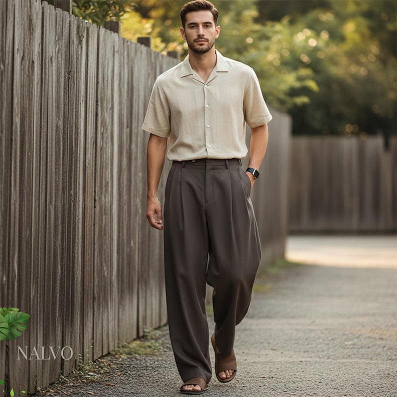 May include: A man wearing a beige short-sleeved button-down shirt and dark brown pleated trousers. He is wearing a black watch and brown sandals. The outfit is suitable for warm weather. The background includes a wooden fence and a paved path.
