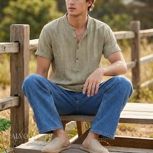 May include: A light green linen shirt with a mandarin collar and short sleeves, worn with blue denim jeans. The outfit is completed with beige slip-on shoes. The person is sitting on a wooden bench outdoors.