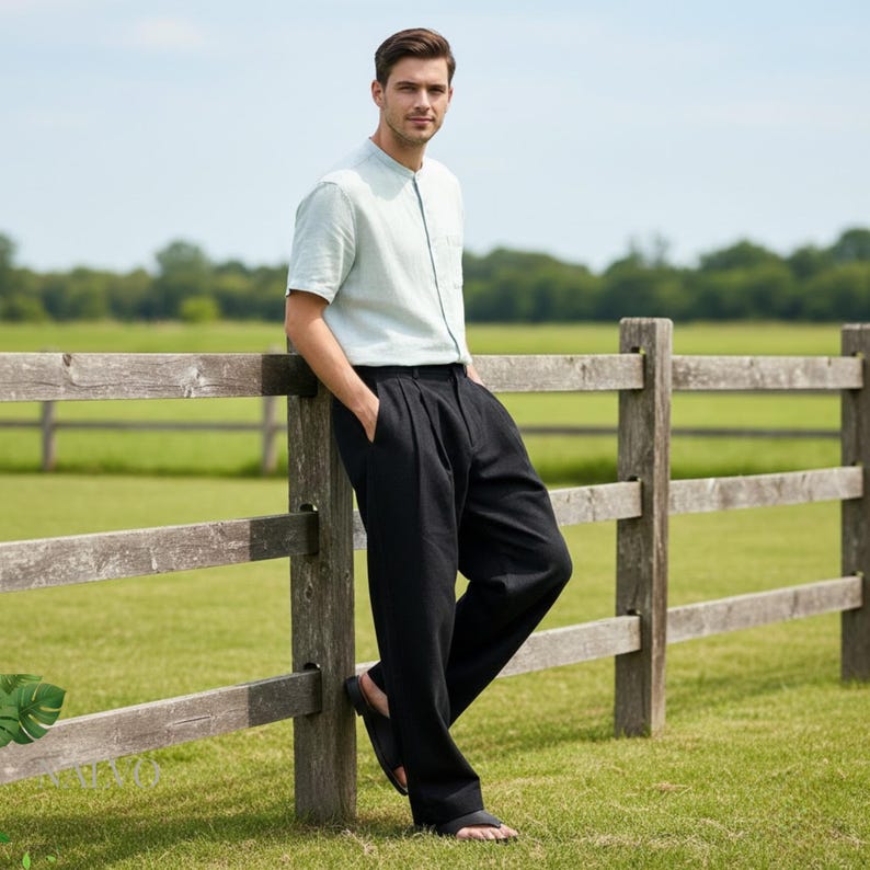 May include: A person wearing a light blue short-sleeved shirt and black pants, leaning against a wooden fence. The pants have a pleated front. The person is also wearing black sandals. The background is a green field and blue sky.