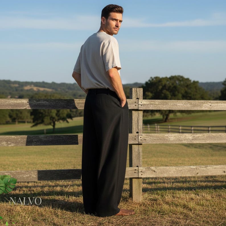 May include: A person wearing a light gray short-sleeve shirt and black wide-leg pants. The person is standing near a wooden fence in a field. The pants have pockets.