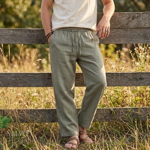 Op de afbeelding: Olijfgroene linnen broek met een trekkoord in de taille. De broek wordt gecombineerd met een crèmekleurig t-shirt en bruine sandalen. Het model leunt tegen een verweerde houten hek in een veld met hoog gras.