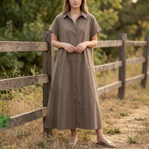 Puede incluir: Vestido largo abotonado de color verde oliva con mangas cortas. El vestido tiene cuello camisero y llega hasta los tobillos. La persona lleva sandalias. Se ve una valla de madera y vegetación en el fondo.