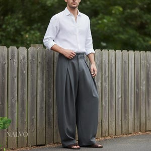 May include: A person wearing a white button-down shirt and gray pleated trousers. The trousers have a wide leg design. The image is taken outdoors with a wooden fence in the background. The brand name "NALVO" is visible on the left side.