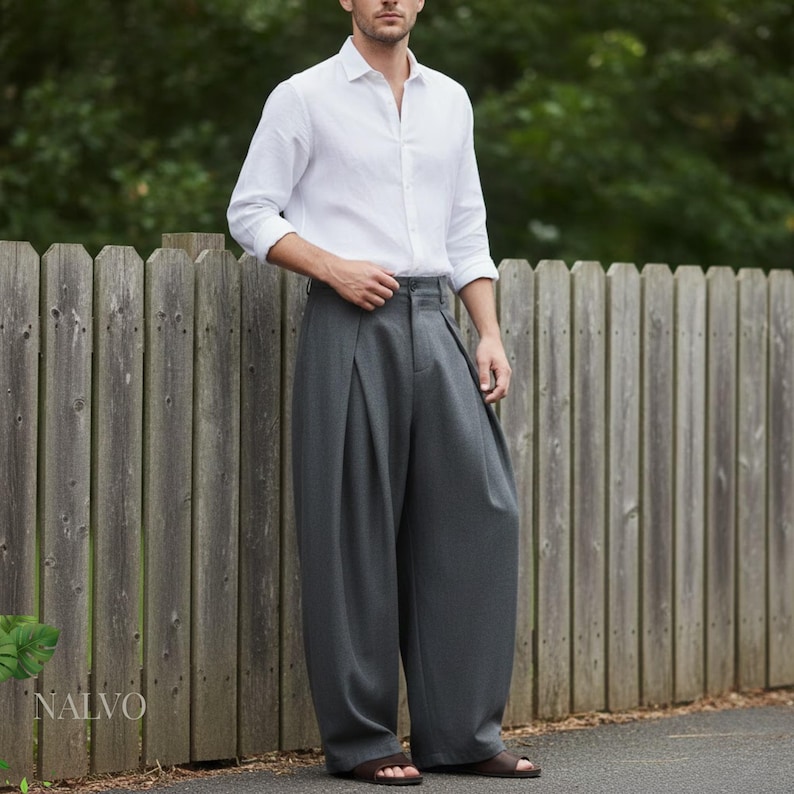 May include: A person wearing a white button-down shirt and gray pleated trousers. The trousers have a wide leg design. The image is taken outdoors with a wooden fence in the background. The brand name "NALVO" is visible on the left side.