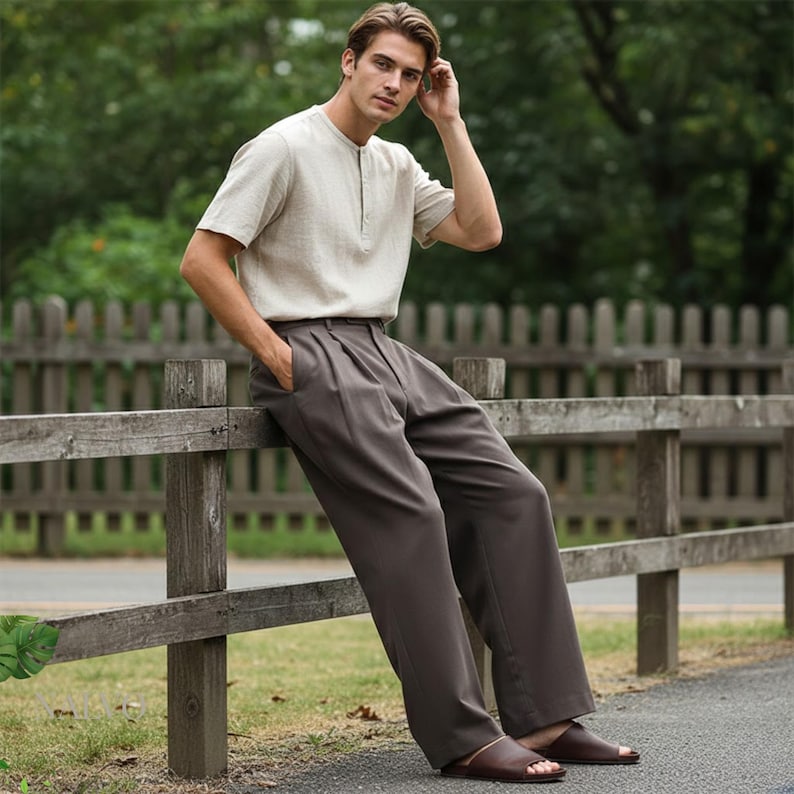 May include: A light beige short-sleeved shirt with a mandarin collar and dark brown trousers. The trousers have a pleated front and a wide leg. The person is wearing brown leather sandals. The outfit is casual and relaxed.