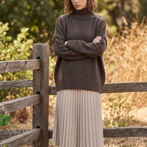 Könnte beinhalten: Ein dunkelbrauner Langarmpullover mit hohem Stehkragen und gerippten Bündchen, kombiniert mit einem hellbeigen, wadenlangen Rock mit vertikalen Falten. Der Pullover hat sichtbare Nahtdetails an den Schultern und Ärmeln.