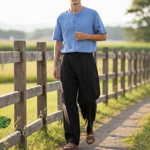 May include: A person wearing a light blue short-sleeve shirt with a mandarin collar and black pleated pants. The person is wearing brown sandals and walking on a gravel path next to a wooden fence.