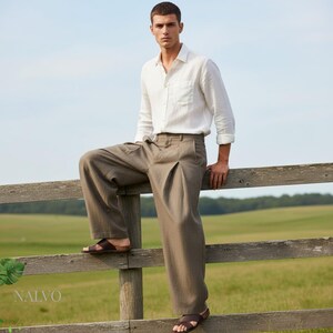 May include: A man wearing a white button-down shirt, brown pleated trousers, and brown leather sandals. The man is leaning on a wooden fence in a field. The brand name "NALVO" is visible in the lower left corner.