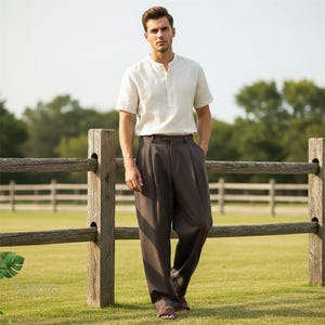 May include: A man wearing a short-sleeved, off-white linen shirt with a mandarin collar and dark brown pleated trousers. The outfit is completed with sandals. The background features a wooden fence and a grassy field.