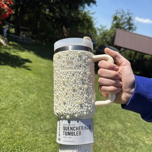 May include: A white Stanley Quencher H2.0 Tumbler, adorned with numerous white pearls. The tumbler has a handle and a lid. The text "QUENCHER H2.0 TUMBLER" is visible on the label. The tumbler holds 40 oz / 1.18L.