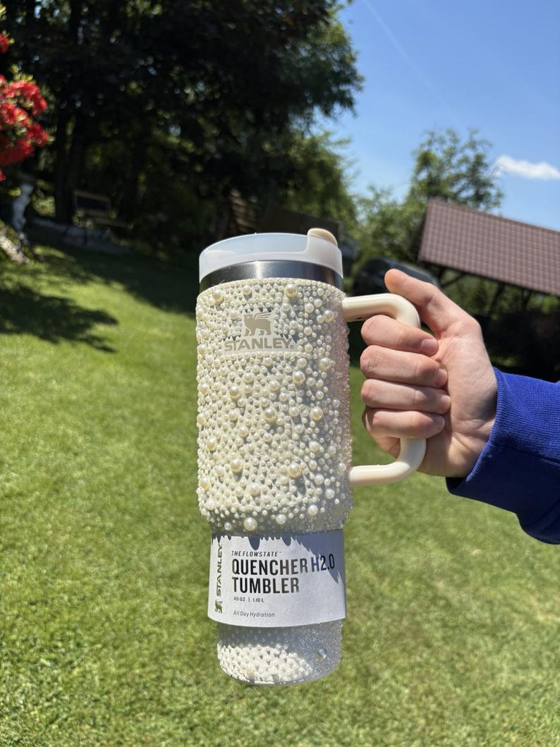 May include: A white Stanley Quencher H2.0 Tumbler, adorned with numerous white pearls. The tumbler has a handle and a lid. The text "QUENCHER H2.0 TUMBLER" is visible on the label. The tumbler holds 40 oz / 1.18L.