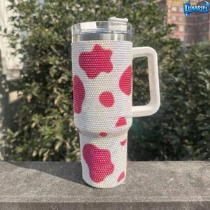 May include: A white tumbler with a handle, covered in small, sparkling rhinestones. The tumbler features a pattern of large, pink cow spots. The lid is silver, and the handle is white. The tumbler is designed to keep drinks cold or hot.