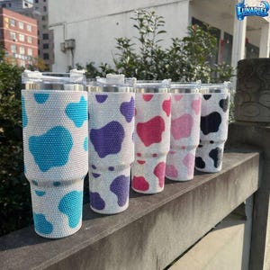 May include: A row of five stainless steel tumblers with clear lids. Each tumbler is covered in white rhinestones and features a cow print design in various colors: blue, purple, red, pink, and black. The tumblers are lined up on a ledge.