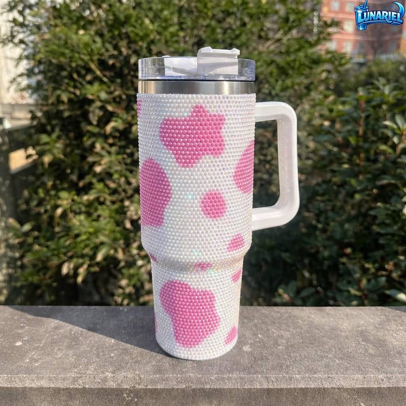 May include: A white and pink cow print tumbler with a handle. The tumbler is covered in small, sparkling rhinestones. The lid is silver and white, and the handle is white.