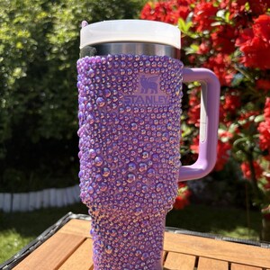 May include: A purple Stanley tumbler covered in iridescent rhinestones. The tumbler has a white lid and a purple handle. The Stanley logo is visible on the front. The tumbler is sitting on a wooden table outdoors.